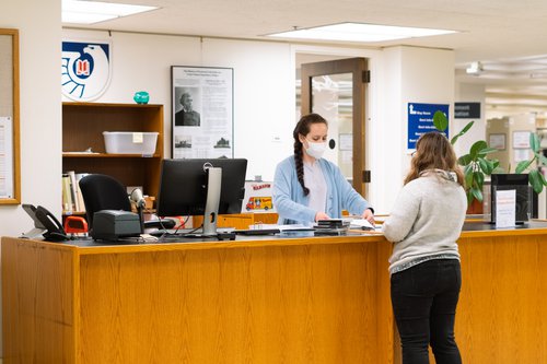 person standing behind wooden counter wearing mask, helping someone in front of counter